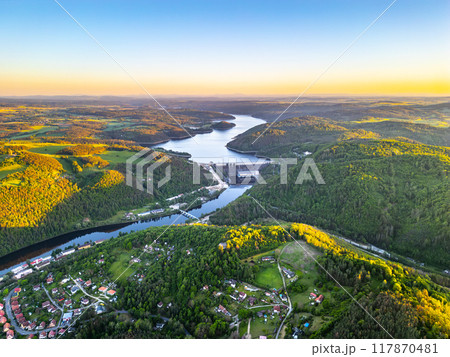 A sunset aerial shot of Orlik Reservoir in Czechia shows a winding river, hills, and a quaint town in the valley with vibrant blue and golden skies. A sunset aerial shot of Orlik Reservoir in Czechia shows a winding river, hills, and a quaint town in the valley with vibrant blue and golden skies. 117870481