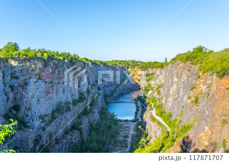 In the bright morning light, the stunning Velka Amerika Quarry showcases its towering cliffs and tranquil blue waters, inviting exploration among lush greenery and vibrant natural scenery. 117871707