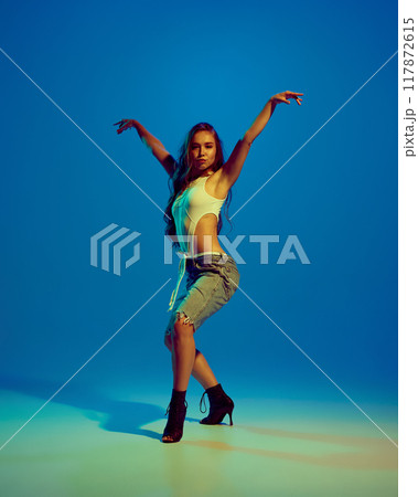 Beautiful young woman in black heels and short performing creative dance against blue studio background in neon light Beautiful young woman in black heels and short performing creative dance against blue studio background in neon light 117872615