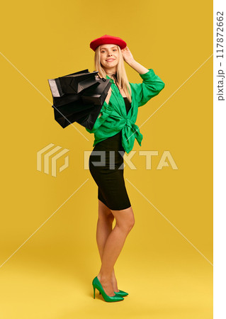 Beowulf young woman in elegant outfit, black dress, red cap and green shirt, standing with shopping bags against yellow studio background Beowulf young woman in elegant outfit, black dress, red cap and green shirt, standing with shopping bags against yellow studio background 117872662
