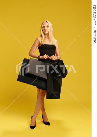 Full-length portrait of beautiful young woman in elegant black dress standing with many shopping bags against yellow studio background Full-length portrait of beautiful young woman in elegant black dress standing with many shopping bags against yellow studio background 117872683