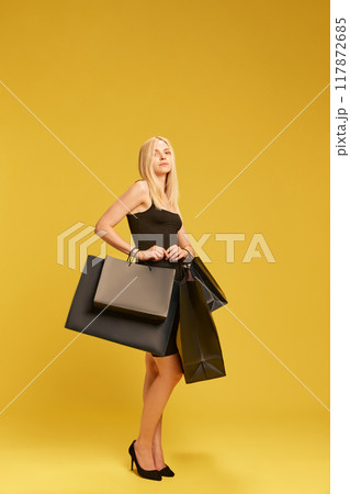 Good shopping deals. Elegant young woman in black dress carrying shopping bags against yellow studio background 117872685