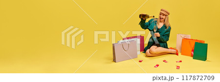 Happy young woman sitting on floor with champagne around colorful shopping bags and sale labels against yellow studio background 117872709