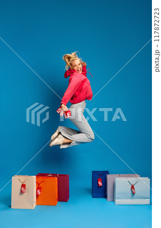 Happy young woman in casual clothes jumping around many colorful shopping bags with percentage signs on it against blue studio background Happy young woman in casual clothes jumping around many colorful shopping bags with percentage signs on it against blue studio background 117872723