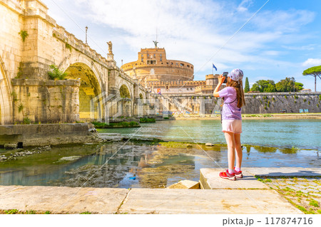 A young girl captures a photo of Castel SantAngelo and a bridge in Rome with her smartphone on a bright sunny day. Rome, Italy 117874716