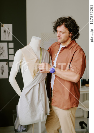 Caucasian curly haired tailor standing next to white plastic mannequin and measuring cotton fabric Caucasian curly haired tailor standing next to white plastic mannequin and measuring cotton fabric 117875265