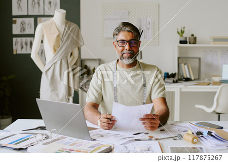Portrait of excited tailor with measurement tape on shoulders holding fresh sketch and looking at camera 117875267