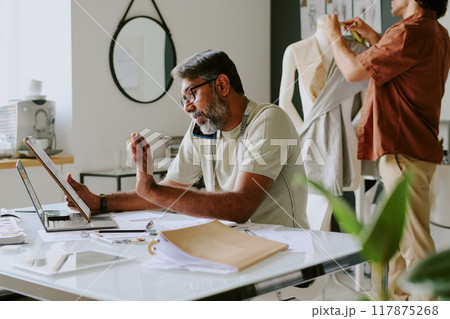 Indian professional designer discussing details with customer via smartphone while tailor creating new dress 117875268