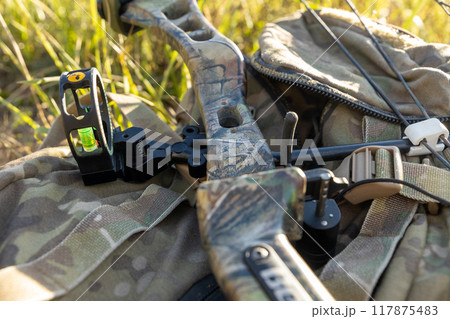 In a sunlit clearing, a camouflaged hunting compound bow rests on the backpack, poised for action In a sunlit clearing, a camouflaged hunting compound bow rests on the backpack, poised for action 117875483