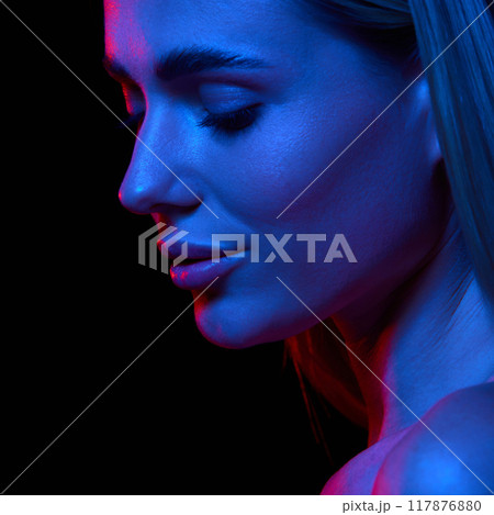 Young beautiful woman with flawless, clear skin showcasing impact of dedicated skincare routine against black studio background in neon light 117876880