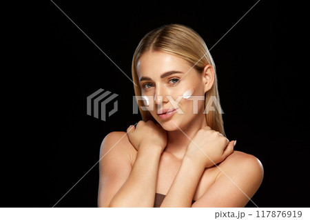 Portrait of attractive young blonde woman with moisturizing cream on cheeks against black background. Skincare routine and advanced cosmetic care. 117876919