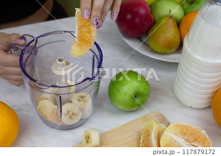Woman hand lowers orange into blender container while making sports cocktail with milk at home. Boosting immunity and vitamins in the body with healthy drink Woman hand lowers orange into blender container while making sports cocktail with milk at home. Boosting immunity and vitamins in the body with healthy drink 117879172