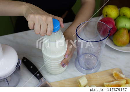 Girl opens bottle of milk to use in blender with fruits while making milkshake. Sports drink with vitamins from fruits and milk, ingredients for healthy drink recipe Girl opens bottle of milk to use in blender with fruits while making milkshake. Sports drink with vitamins from fruits and milk, ingredients for healthy drink recipe 117879177