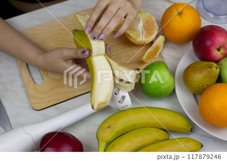 Female hands peeling banana on kitchen table with fruits on background. Girl preparing sports fruit cocktail. Healthy nutrition and lifestyle at home 117879246
