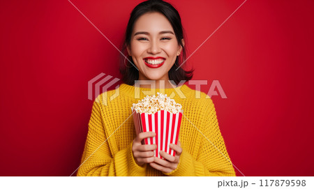 Cheerful young asian woman in yellow sweater enjoying popcorn from cinema box 117879598