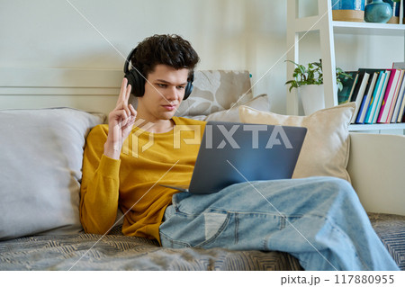 Young male college student in headphones sitting on couch, talking in web camera of laptop Young male college student in headphones sitting on couch, talking in web camera of laptop 117880955