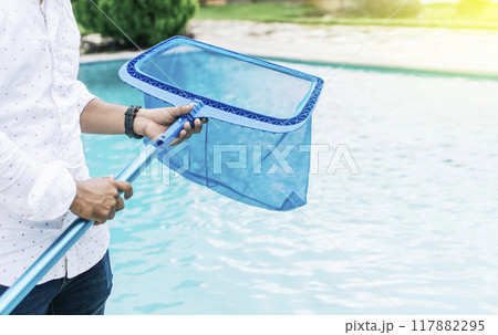 Hands holding a skimmer with blue pool in the background. A man cleaning pool with leaf skimmer. Man cleaning the pool with the Skimmer, Person with skimmer cleaning pool 117882295