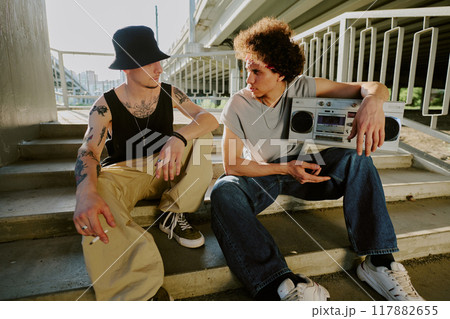 Biracial bros chatting and sitting on stairs while smoking and listening to music Biracial bros chatting and sitting on stairs while smoking and listening to music 117882655