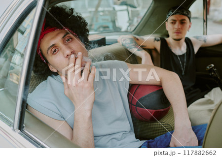 Curly haired man looking at camera while smoking, his tattooed friend looking at him 117882662