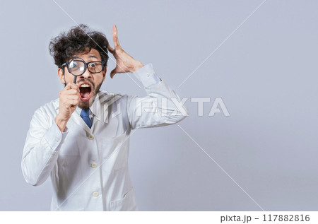 Surprised male scientist in lab coat looking at camera with magnifying glass. Mad scientist holding a magnifying glass isolated, Man in white coat with magnifying glass looking at camera 117882816