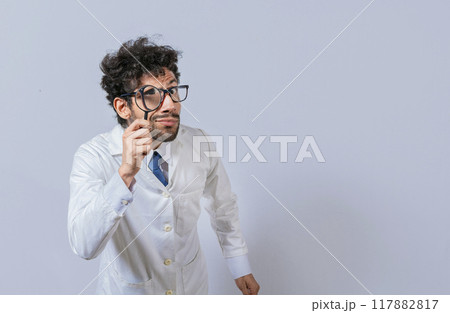 Mad scientist holding a magnifying glass isolated, Man in white coat with magnifying glass looking at camera, Surprised male scientist in lab coat looking at camera with magnifying glass 117882817