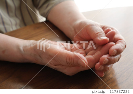 机上で両手を重ねる高齢女性の手元 117887222