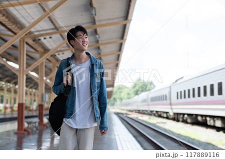 A young man carries a backpack and waits for the train at the train station to travel. 117889116