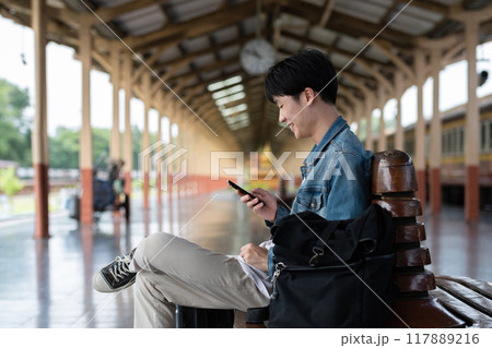 Young man with backpack checking social media on phone waiting for train at train station to travel 117889216