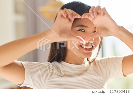 Smiling Woman Framing Her Eye With Heart-Shaped Hands In Bright Room Smiling Woman Framing Her Eye With Heart-Shaped Hands In Bright Room 117893418