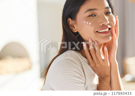 Healthy Skin Routine. Smiling Woman Applying Skincare Cream in Bright Natural Light Indoors 117893433