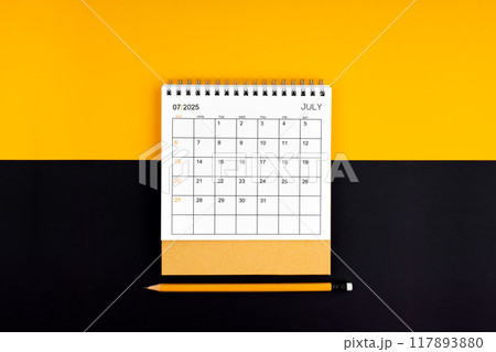 Top view of July 2025 desk calendar and pencil on yellow and black background. 117893880
