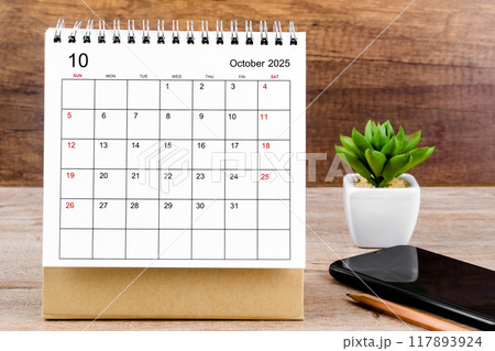 October 2025 desk calendar on wooden table with black smartphone. 117893924