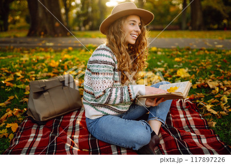 Beautiful curly woman in hat on mat with book in autumn park. Relaxation, solitude with nature. 117897226