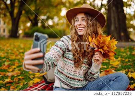 Selfie time. Beautiful woman takes selfie on smartphone in hat on meadow in autumn park on plaid. Selfie time. Beautiful woman takes selfie on smartphone in hat on meadow in autumn park on plaid. 117897229