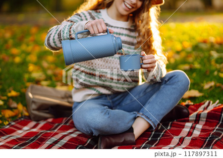 Curly woman with thermos in clearing among fallen leaves in autumn park, enjoying hot drink. 117897311