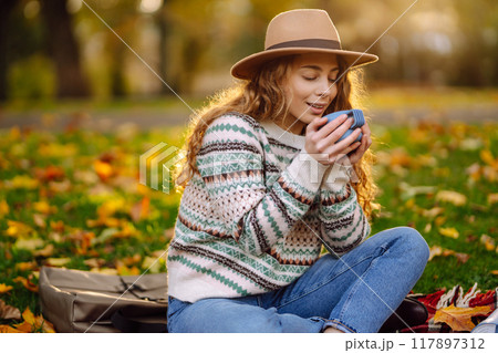 Curly woman with thermos in clearing among fallen leaves in autumn park, enjoying hot drink. Curly woman with thermos in clearing among fallen leaves in autumn park, enjoying hot drink. 117897312