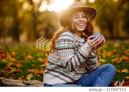 Curly woman with thermos in clearing among fallen leaves in autumn park, enjoying hot drink. Curly woman with thermos in clearing among fallen leaves in autumn park, enjoying hot drink. 117897313