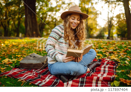 Beautiful curly woman in hat on mat with book in autumn park. Relaxation, solitude with nature. Beautiful curly woman in hat on mat with book in autumn park. Relaxation, solitude with nature. 117897404