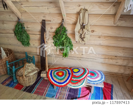 A room with a wooden log wall on which a two-handed saw rests, birch brooms hang, an old backpack. There is also an old wool spinning wheel with round mini carpets with national Tatar patterns A room with a wooden log wall on which a two-handed saw rests, birch brooms hang, an old backpack. There is also an old wool spinning wheel with round mini carpets with national Tatar patterns 117897413