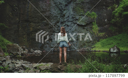 Tourist woman enjoy jungle waterfall landscape standing on rock stone. Girl relax outdoor lifestyle travel on summer holiday vacation. Picturesque scene panoramic shot. Dramatic dark toning, back view Tourist woman enjoy jungle waterfall landscape standing on rock stone. Girl relax outdoor lifestyle travel on summer holiday vacation. Picturesque scene panoramic shot. Dramatic dark toning, back view 117897799
