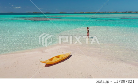 Woman enjoying a tropical beach vacation, walking on a sandbar with a kayak nearby, surrounded by turquoise ocean and blue sky on a sunny day 117897821