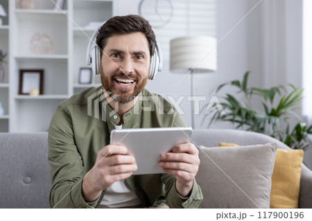 Smiling man wearing headphones holding tablet while sitting on sofa in brightly lit living room. Person enjoying digital content, technology, and relaxation during leisure time. Smiling man wearing headphones holding tablet while sitting on sofa in brightly lit living room. Person enjoying digital content, technology, and relaxation during leisure time. 117900196