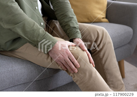 Middle-aged man holding knee in discomfort while sitting on sofa. Concept of joint pain, aging, health issues. Image captures emotion of concern regarding personal wellness and mobility challenges. 117900229