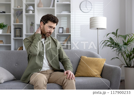 Man experiencing neck pain while sitting on couch at home. Wearing green shirt and beige pants, hand on neck. Room interior includes sofa, pillows, plant, shelf, and lamp. 117900233