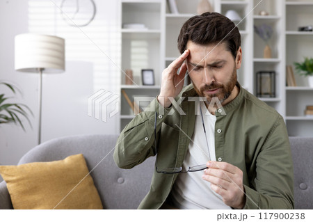 Man sitting on gray couch experiencing headache, holding glasses with pained expression. Shows concept of stress, frustration, fatigue in home environment. 117900238