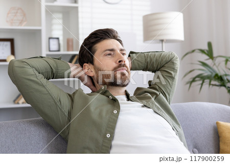 Relaxed man sits on couch with hands behind head, reflecting in modern living room. Casual attire complements calm demeanor, conveying tranquility and peacefulness. Relaxation and contemplation 117900259