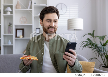 Smiling man using smartphone while enjoying pizza in cozy living room. Wearing casual green shirt, feeling relaxed, engaged with technology. Concept of leisure, technology, comfort. 117900267