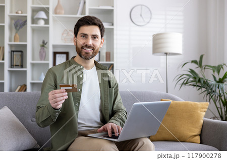 Confident man sitting on sofa holding credit card and using laptop for online shopping. Shows convenience and enjoyment of digital transactions at home. Ideal for themes of modern lifestyle Confident man sitting on sofa holding credit card and using laptop for online shopping. Shows convenience and enjoyment of digital transactions at home. Ideal for themes of modern lifestyle 117900278