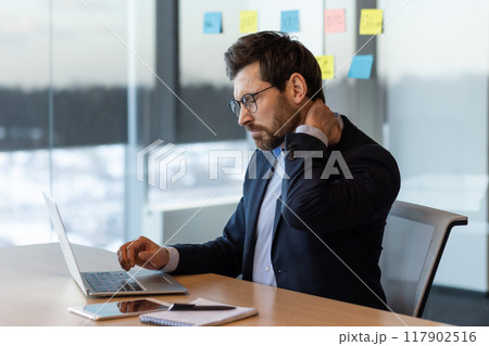 Businessman in suit sitting at desk using laptop, appearing uncomfortable due to neck pain. Office with focused work atmosphere illustrates concept of work-related stress and physical discomfort 117902516