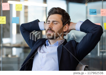 Businessman relaxing at office desk with hands behind head, eyes closed. Enjoying moment of peace and calm, taking break from work, wearing a formal suit. Businessman relaxing at office desk with hands behind head, eyes closed. Enjoying moment of peace and calm, taking break from work, wearing a formal suit. 117902536